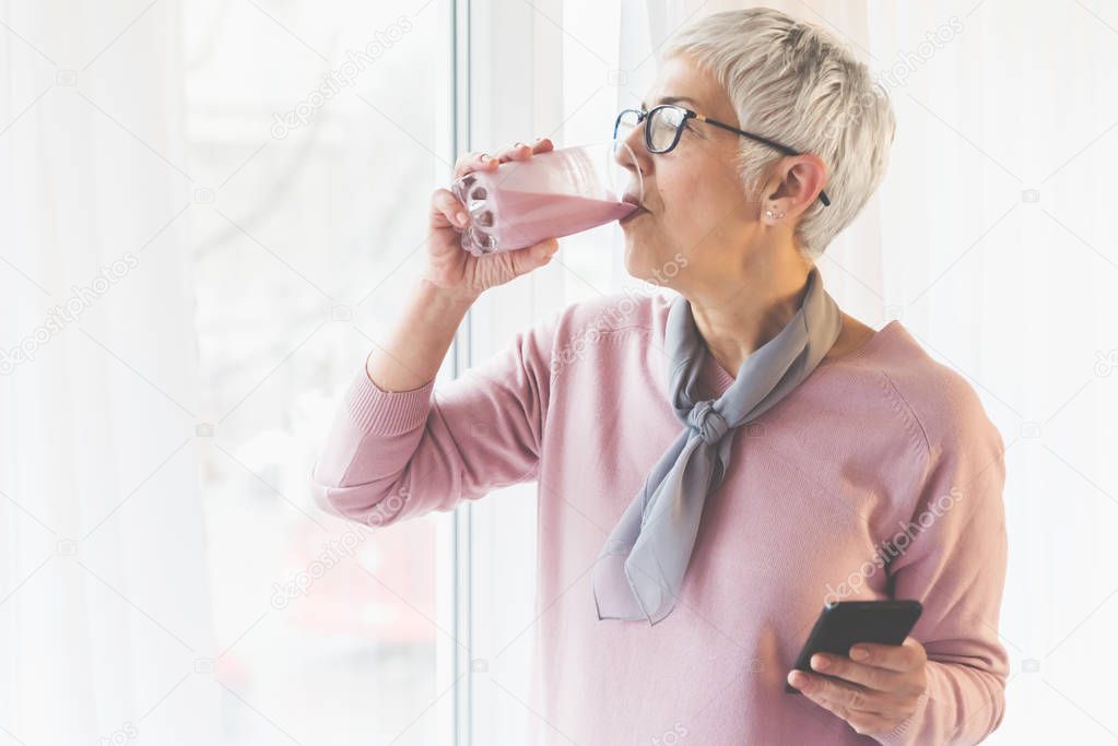 senior woman with modern short gray hair and glasses drinking healthy fresh fruit smoothie by window and holding mobile phone