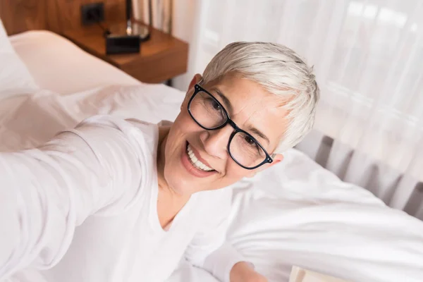 有吸引力的成熟女人与现代眼镜和美丽的微笑采取自拍在她的明亮的现代卧室 — 图库照片