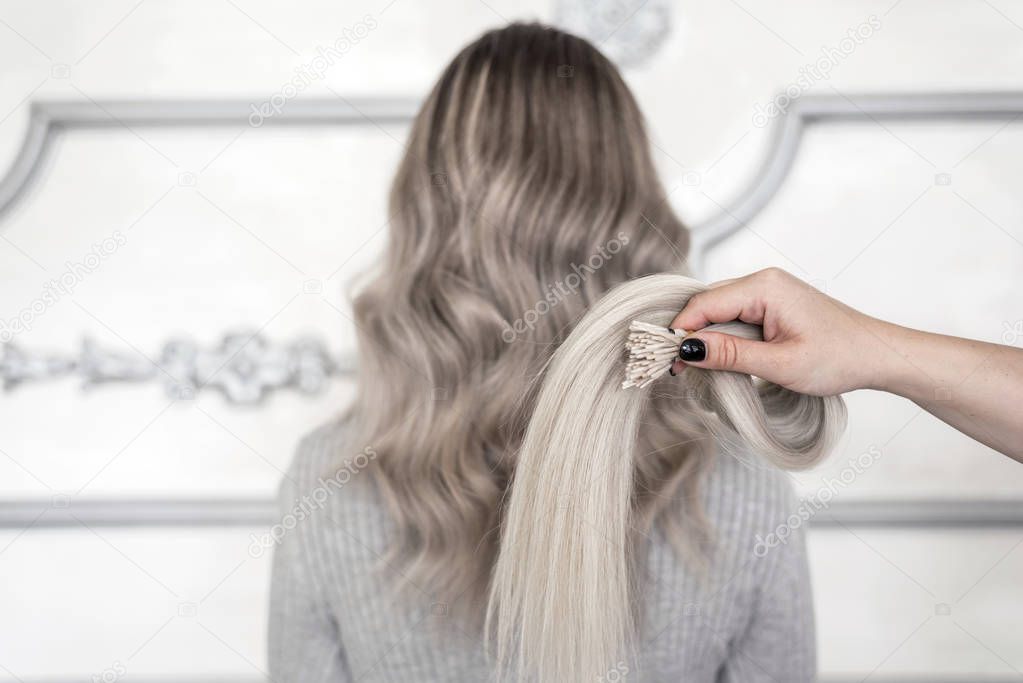 Female hand holding sample of hair extension and in background standing girl with blond ombre hairstyle