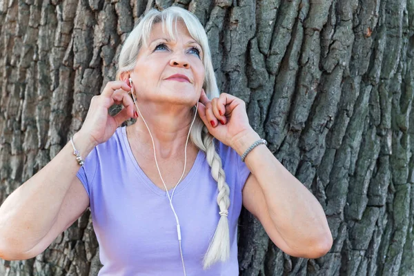 シニア女性置くヘッドセットのために耳に音楽を聴くために公園 — ストック写真