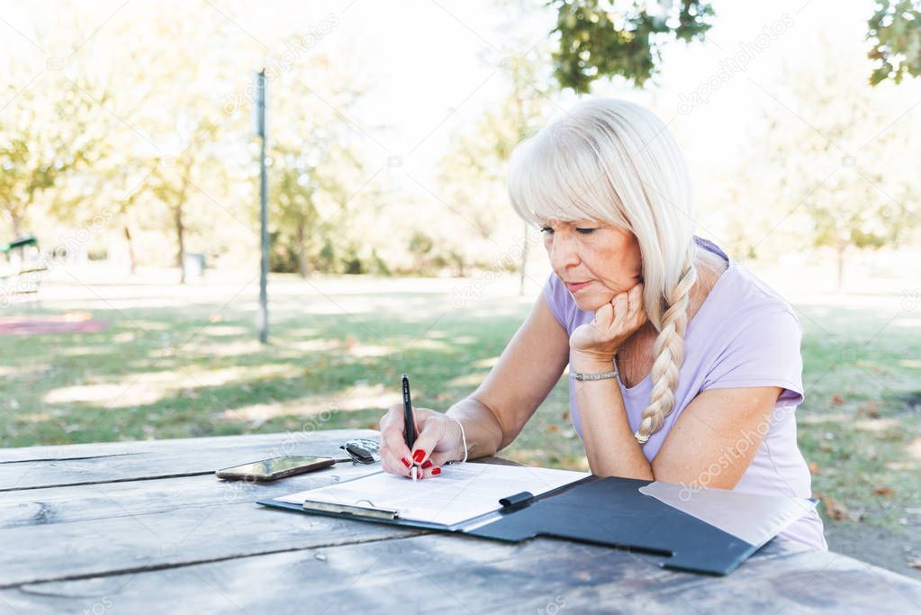 Serious senior woman with long natural hair sitting at park making notes in her diary