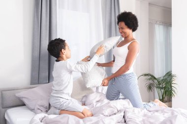 Oyuncu anne ve oğul yatakta yastıkla kavga ediyor.