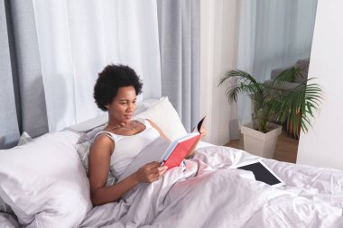 Afro-Amerikalı bir kadın evde yatakta kitap okuyor.