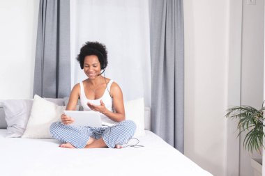 Afro-Amerikan kadın video sohbetleri yatakta dijital tablet ve kulaklık kullanıyor.