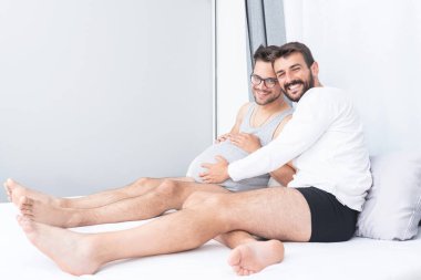 Mutlu eşcinsel adam hamile erkek arkadaşının karnına dokunuyor evde yatakta oturuyor.
