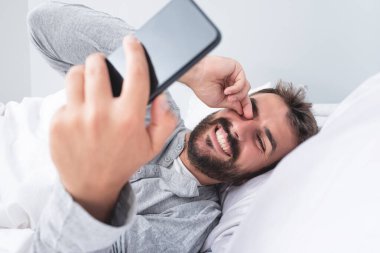 Adam evde yatakta dinlenirken cep telefonu kullanırken eğleniyor.