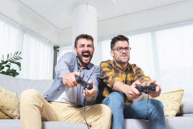 Evdeki kanepede oturmuş video oyunu oynayan iki heyecanlı erkek arkadaş.