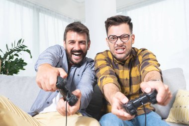 İki genç erkek arkadaş oturma odasında agresif bir şekilde video oyunu oynuyor.