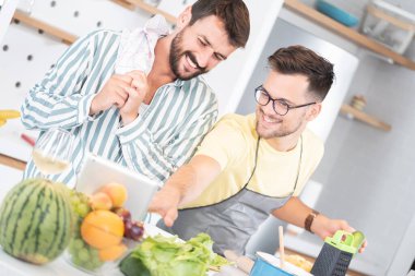 İki mutlu erkek arkadaş mutfakta yemek pişirirken dijital tablet kullanarak yemek tarifi öğretiyor.