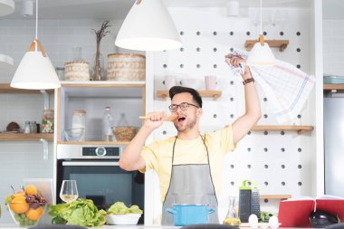 Mutfakta yemek pişirirken elinde spatulayla şarkı söyleyen genç adam.