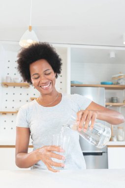 Afro-Amerikalı kadın mutfaktaki bardağa mutlulukla su dolduruyor.