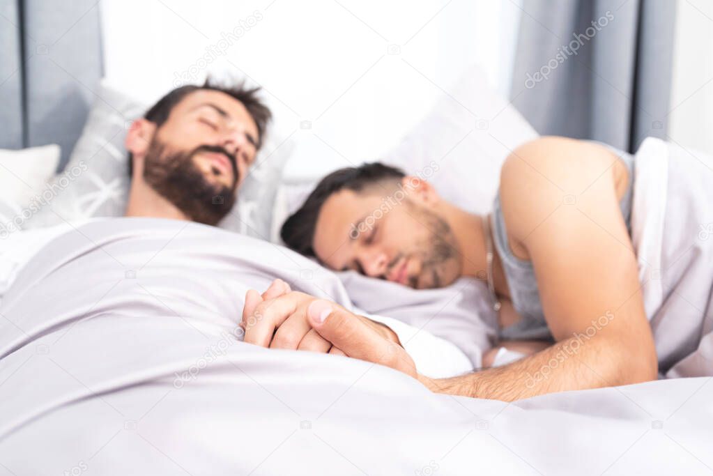 Two gay men sleeping in bed with hands clasped at home