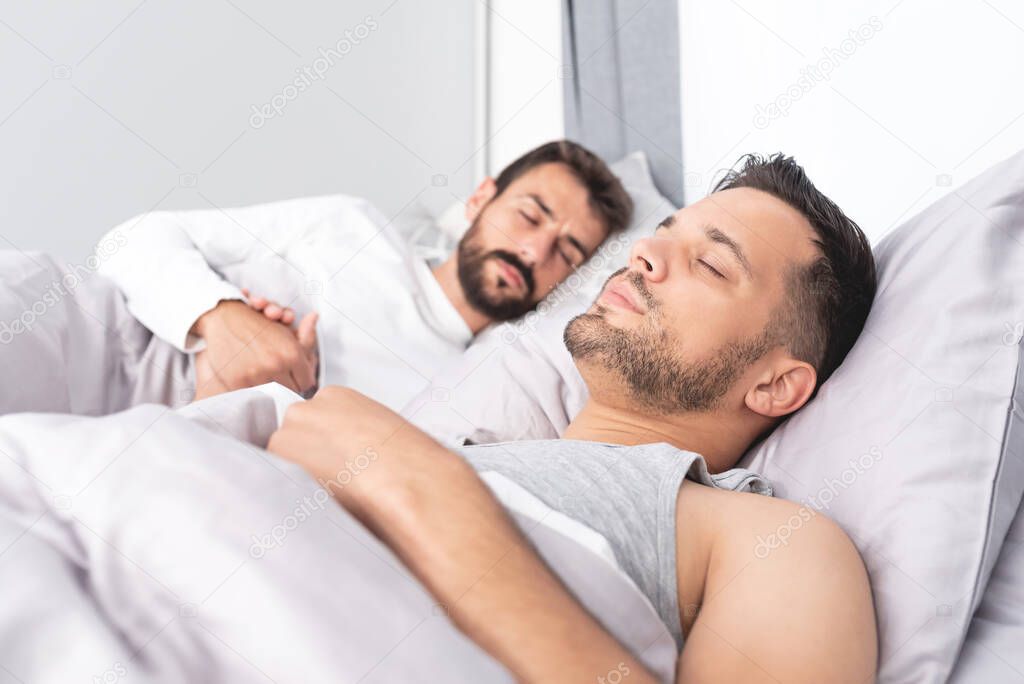 Young gay couple sleeping on bed with hands clasped at home