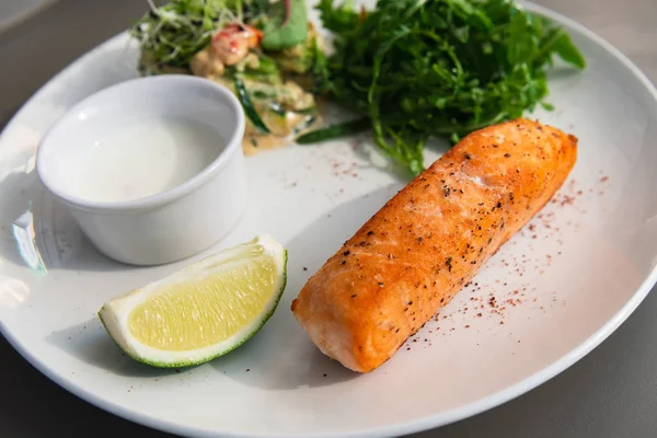 Grilled salmon steak on the side with arugula and salad. Served with a sauce and lime on a white plate. White sauce.