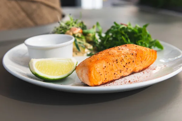 Grilled salmon steak on the side with arugula and salad. Served with a sauce and lime on a white plate in the restaurant. Closeup.