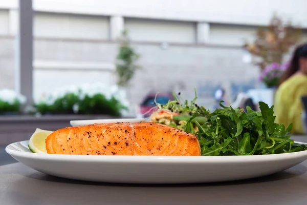 Grilled salmon steak on the side with arugula and salad. Served with a sauce and lime on a white plate in the restaurant.