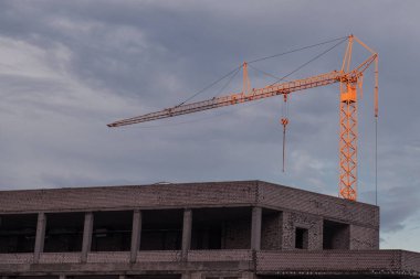 Akşam bulutlu gökyüzüne karşı yapım aşamasında olan bina. İş sırasında üst kısmında büyük turuncu vinç. Alt görünüm. Günbatımı.