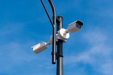 Mavi gökyüzüne karşı metal lamba direğine ait iki beyaz güvenlik kamerası. Tüm bölgenin güvenliği. Güvenlik servisi izliyor. Kopyalama alanı.