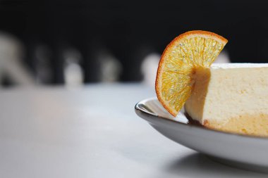 Kurutulmuş portakal parçasıyla süslenmiş bir dilim cheesecake. Lokantada beyaz masada servis edilen tatlı. Metin için kopyalama alanı ile kapat.