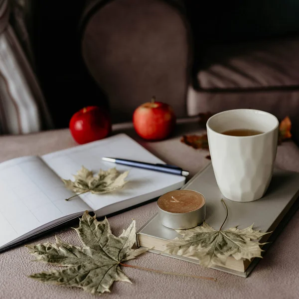 cup of tea with a book in the interior with autumn leaf, notebook and red apples hygge concept