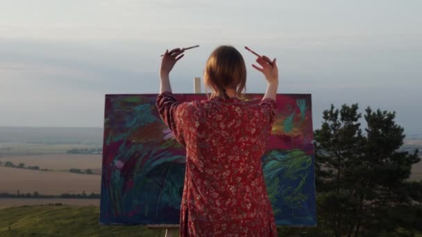 Imagen romántica de la joven artista que pinta en la naturaleza. Chica se para cerca de la foto con cepillos en sus manos y los mueve. — Vídeos de Stock