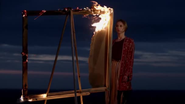 Młoda artystka stoi w naturze o zmierzchu przed sztalugą z płonącym obrazem. — Wideo stockowe