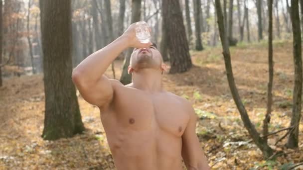 Młody sportowiec z nagim tułowiem stoi w lesie i pije wodę. — Wideo stockowe