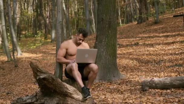 Sterke naakte romp man zit in een herfst park met laptop in zijn handen — Stockvideo