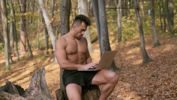 Silny nagi facet tułów siedzi w jesiennym parku z laptopem w rękach — Wideo stockowe