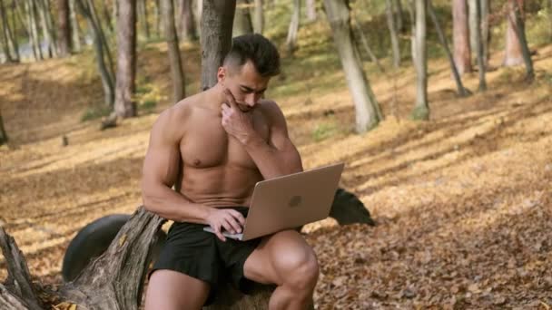 Silny nagi facet tułów siedzi w jesiennym parku z laptopem w rękach. — Wideo stockowe