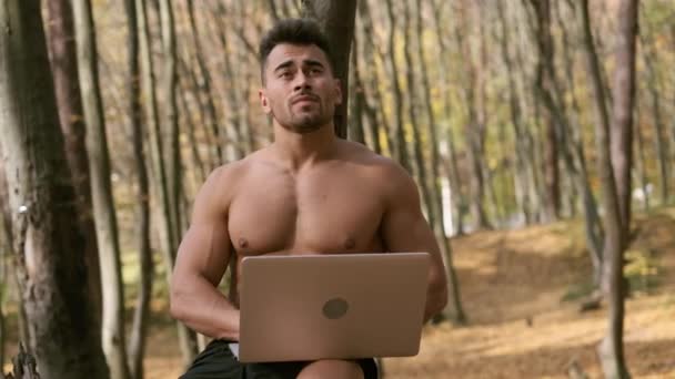 Сильный голый парень сидит в осеннем парке с ноутбуком в руках — стоковое видео