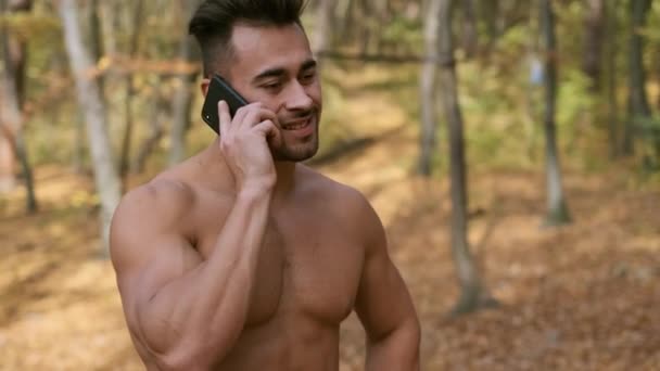 Silny facet z nagim tułowia stoi w jesiennym parku i rozmawia przez telefon komórkowy — Wideo stockowe