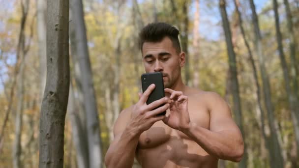 Silny facet z nagim tułowia stoi w jesiennym parku i rozmawia przez telefon komórkowy — Wideo stockowe