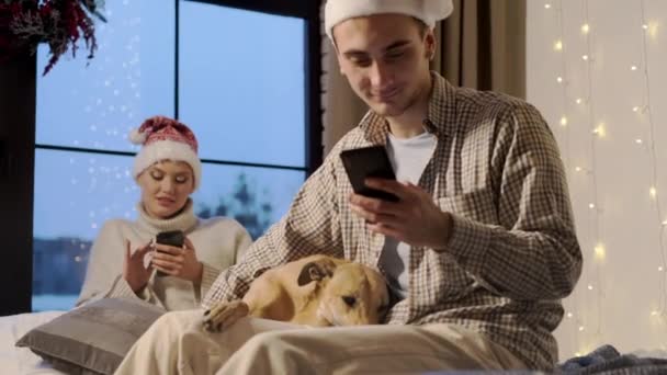新年。若いカップルでサンタは彼らの犬と一緒に家で大晦日の帽子. — ストック動画