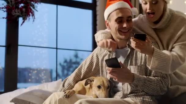 Новый год. Молодая пара в шляпах Санта-Клауса в канун Нового года дома со своей собакой. — стоковое видео