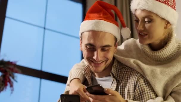 Neujahr. Junges Paar in Nikolausmützen mit Handys in der Hand. — Stockvideo
