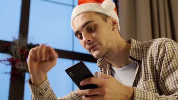 Nouvel An. Le gars au chapeau de Noël Santa regarde dans le téléphone portable. Il bâille. — Video