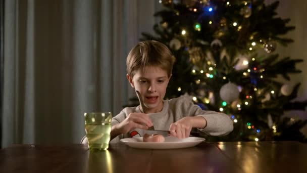 Neujahr. Lustiger Junge sitzt am Tisch und schneidet Wurst auf Teller. Im Hintergrund steht der Weihnachtsbaum — Stockvideo