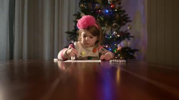 Año Nuevo. Pequeña niña bonita se sienta en la mesa y se basa en un pedazo de papel de dibujo. — Vídeo de stock