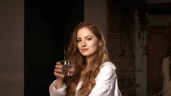 Punatukkainen tyttö valkoisessa paidassa pitää lasia vettä asunnossaan. — kuvapankkivalokuva