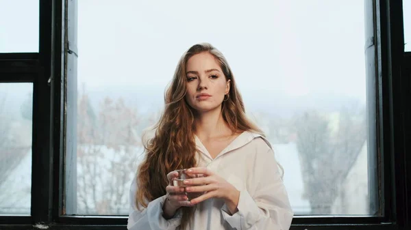 Junges Mädchen mit langen Haaren steht mit Glas Wasser am Fenster. — Stockfoto