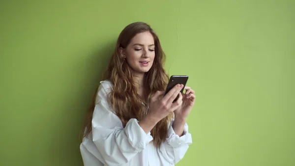 Girl With Long Hair Stands Against Light Green Wall With Mobile Phone In Her Hands.