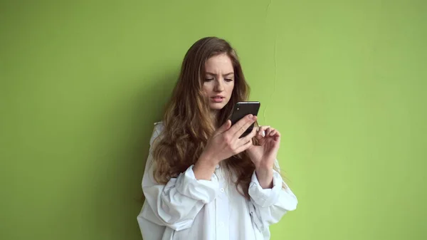 Tyttö pitkät hiukset seisoo vastaan valo vihreä seinä matkapuhelin käsissään. — kuvapankkivalokuva