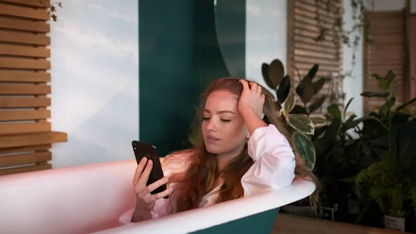 Pen jente med langt hår i hvite gensere setter seg i badekaret på badet.. – stockfoto
