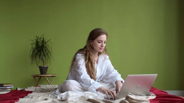 Nuori tyttö käyttää kannettavaa tietokonetta, kun istuu sängyssä asunnossa. — kuvapankkivalokuva