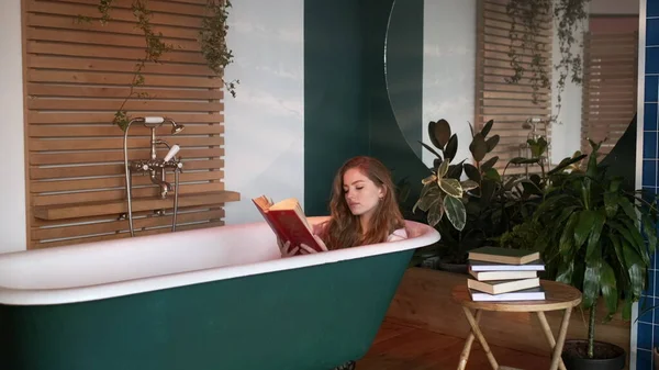 Tyttö istuu kylpyammeessa ilman vettä ja lukemat kirja. tekijänoikeusvapaita kuvapankkikuvia