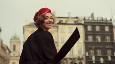 Kırmızı bereli ve kırmızı elbiseli gülümseyen genç kadın şehir meydanında yürüyor..