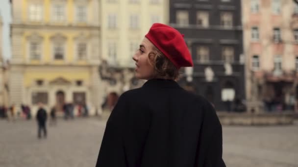 Junge Frau in roter Baskenmütze läuft lächelnd über den Stadtplatz. — Stockvideo