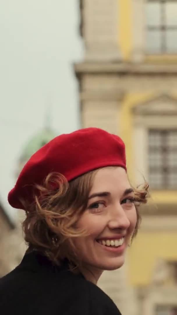 Mujer joven sonriente en boina roja y vestido rojo caminando en la plaza de la ciudad. — Vídeo de stock