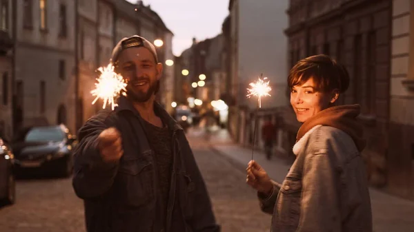 Young Stylish Couple Walking Through The City Street. In Their Hands The Burning Sparklers.